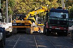 Gelber Bagger auf einer Straße mit Schienen, der in einen grünen Container LKW etwas reinlegt