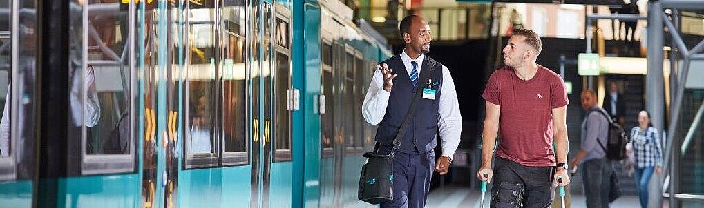 VGF-Mitarbeiter mit einem Fahrgast auf Krücken neben einer VGF-U-Bahn