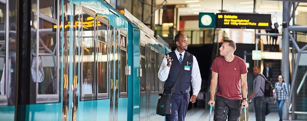VGF-Mitarbeiter mit einem Fahrgast auf Krücken neben einer VGF-U-Bahn