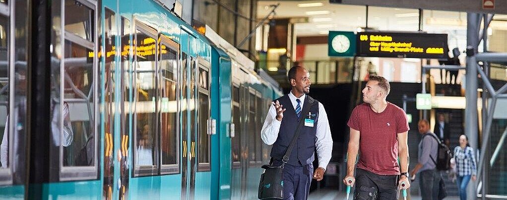 VGF-Mitarbeiter mit einem Fahrgast auf Krücken neben einer VGF-U-Bahn