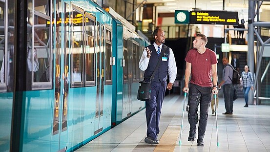 VGF-Mitarbeiter mit einem Fahrgast auf Krücken neben einer VGF-U-Bahn