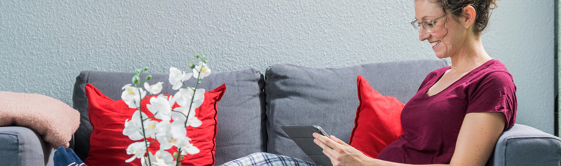 Frau mit Brille und rotem Shirt auf grauer Couch lächelnd mit Tablet in den Händen