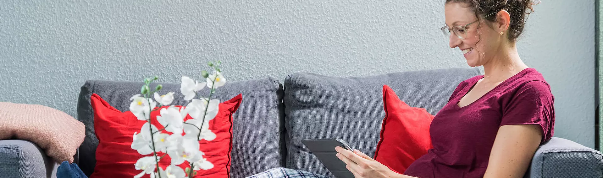 Frau mit Brille und rotem Shirt auf grauer Couch lächelnd mit Tablet in den Händen