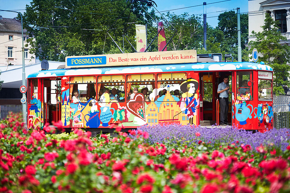 Das Bild zeigt ein Ebbelwei-Expreß-Fahrzeug am Zoo. Im Vordergrund sind Blumen zu sehen.