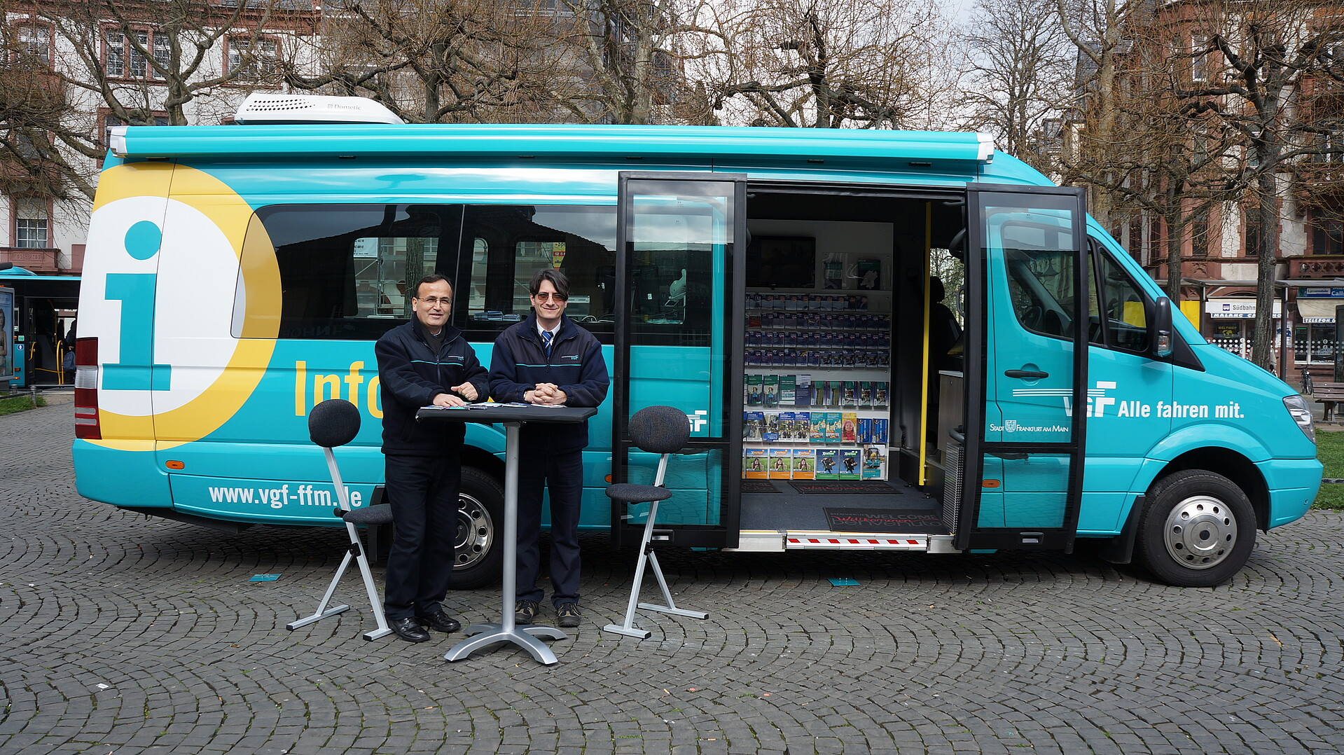 Das Bild zeigt zwei Mitarbeitende der VGF an einem Tisch vor dem Infobus der VGF. 